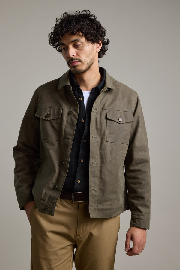 A man with curly dark hair wears the Barkers Benson Waxed Cotton Jacket over a black shirt and tan pants. He stands against a plain background, one hand in his pocket, looking slightly downward with a neutral expression.