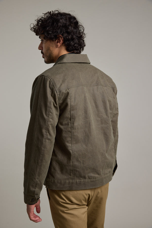 A man with curly dark hair is seen from behind, wearing a Barkers Benson Waxed Cotton Jacket in olive green and khaki pants, standing against a plain light gray background.