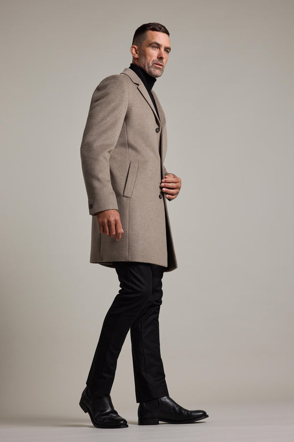 A man with short hair and a beard wears the Morrison Coat by Barkers, a beige wool blend classic. Styled with a black turtleneck, trousers, and dress shoes, he stands in profile against a plain light background, looking ahead confidently.