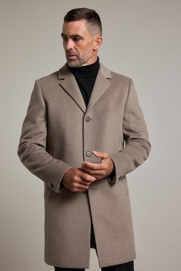 A man with short hair and a beard wears the Barkers Morrison Coat, a taupe wool blend, over a black turtleneck, looking slightly to his left as he adjusts his coat against a plain light background.
