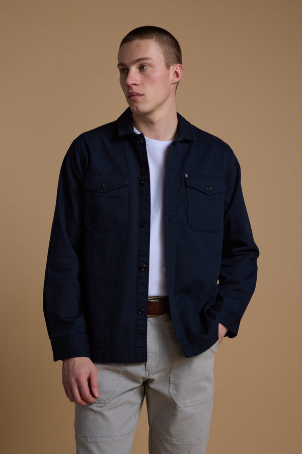 A young man wears the Barkers Gifford Herringbone Jacket over a white t-shirt and light gray pants, standing against a plain beige background and looking slightly to his left with one hand in his pocket.