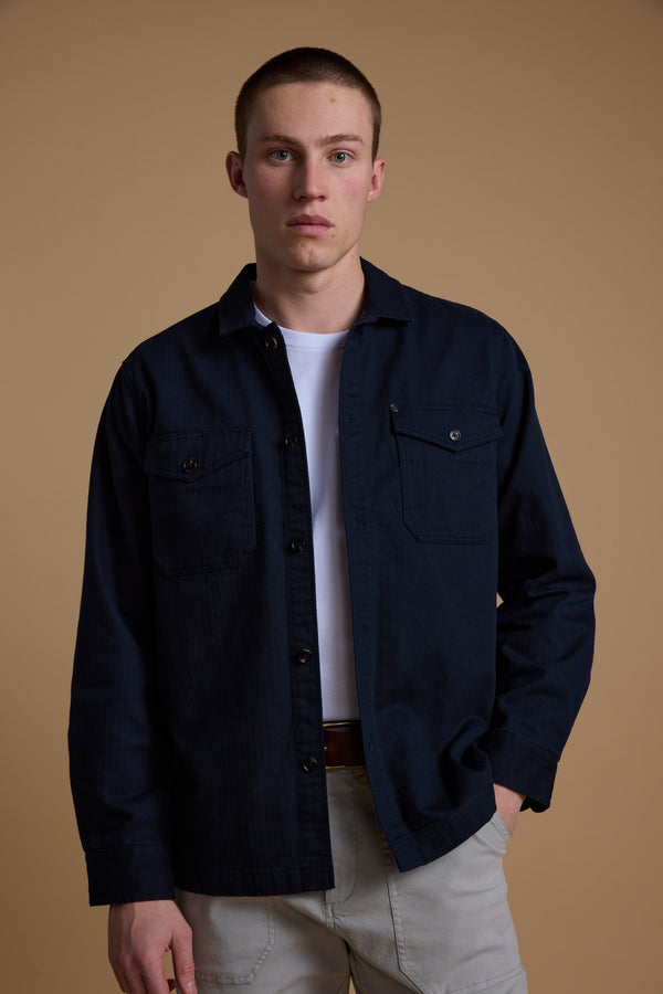 A young man with short hair wears Barkers’ Gifford Herringbone Jacket in dark blue over a white T-shirt and light pants, standing before a plain beige background and looking directly at the camera.