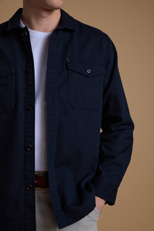 A person wearing the Barkers Gifford Herringbone Jacket in navy over a white t-shirt, paired with light pants and a brown belt, stands against a tan background. The individual's face is partially out of frame.