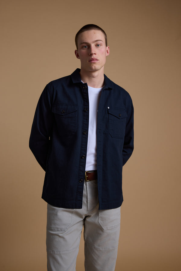 A young man stands against a plain beige background, wearing the Barkers Gifford Herringbone Jacket in navy blue over a white t-shirt and light gray pants, hands behind his back, looking directly at the camera with a neutral expression.
