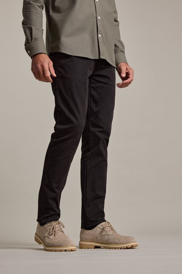 A person wears Barkers Carnaby Chino in black and a light olive button-up shirt, standing against a plain background. The cropped image highlights the lower body and the chinos, made from responsibly sourced cotton.