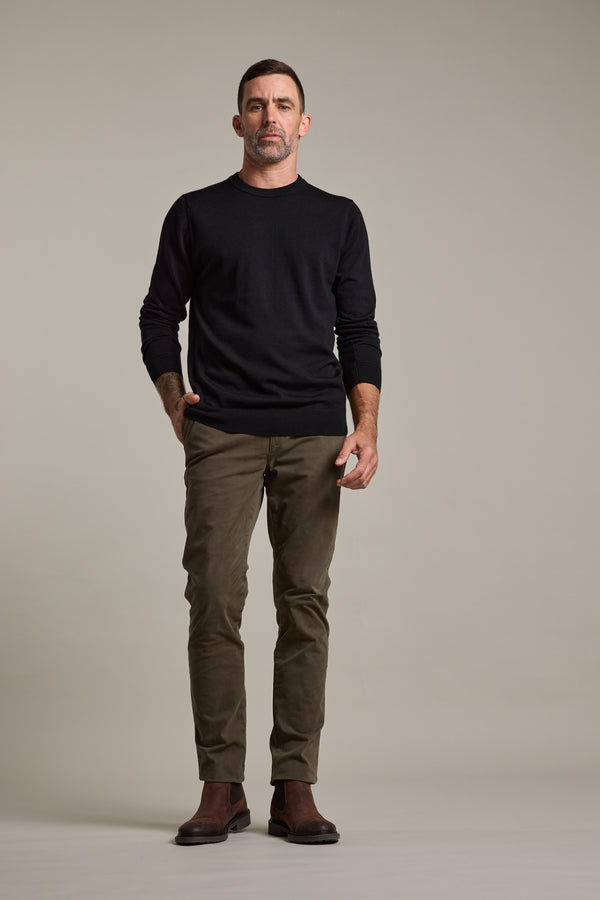 A man stands facing forward against a plain background, wearing a black long-sleeve shirt, Barkers Carnaby Chino in olive green, crafted from responsibly sourced cotton, and brown boots, with one hand in his pocket.