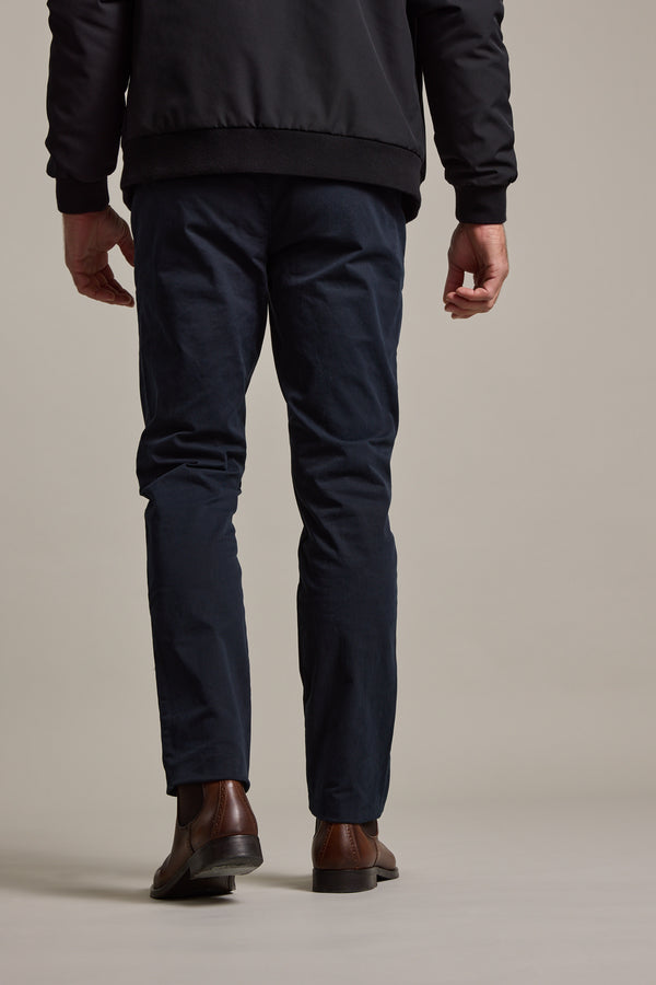 A man faces away from the camera, wearing Barkers' Carnaby Chino in dark blue with a black jacket and brown dress shoes, set against a neutral beige background.
