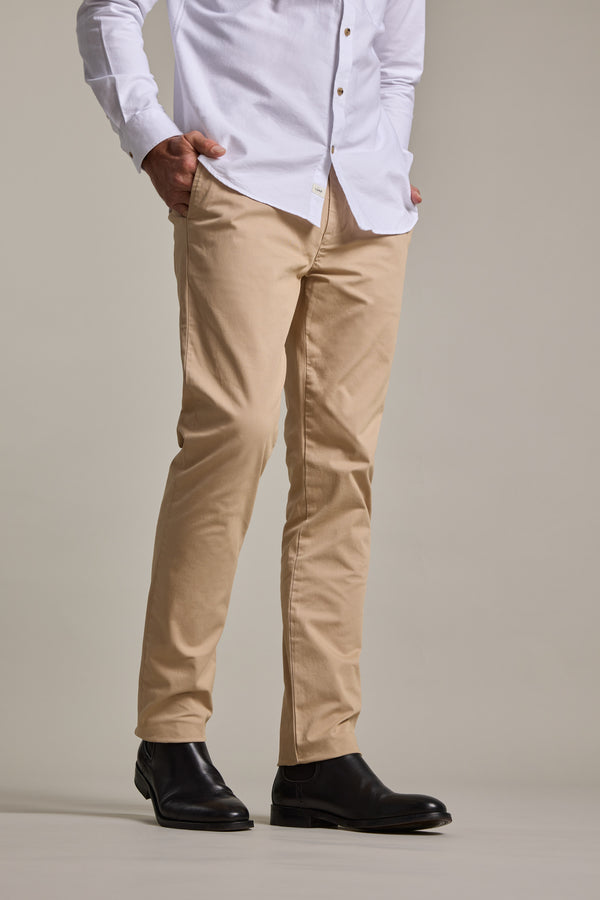 A person stands with hands in pockets against a plain background, wearing a white button-down shirt, Barkers Carnaby Chino men's stretch chinos made from responsibly sourced cotton, and black leather boots. The person's head is not visible in the image.