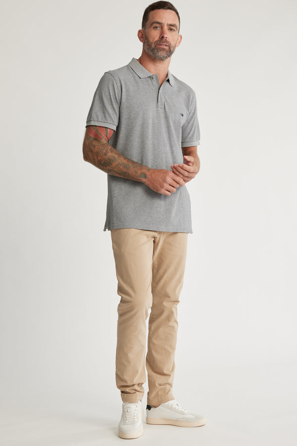 A man with short hair, a beard, and a tattoo sleeve on his left arm stands against a white background wearing a grey polo shirt, white sneakers, and Barkers Beauford Chino O men’s pants made from a cotton elastane blend.