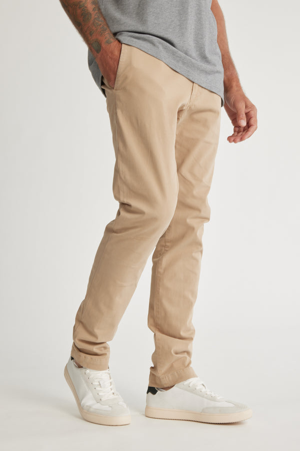 A person in Barkers Beauford Chino O men's pants, made from a cotton elastane blend, with a grey t-shirt and white sneakers stands with one hand in their pocket. The image is cropped from shoulders to feet on a plain background.