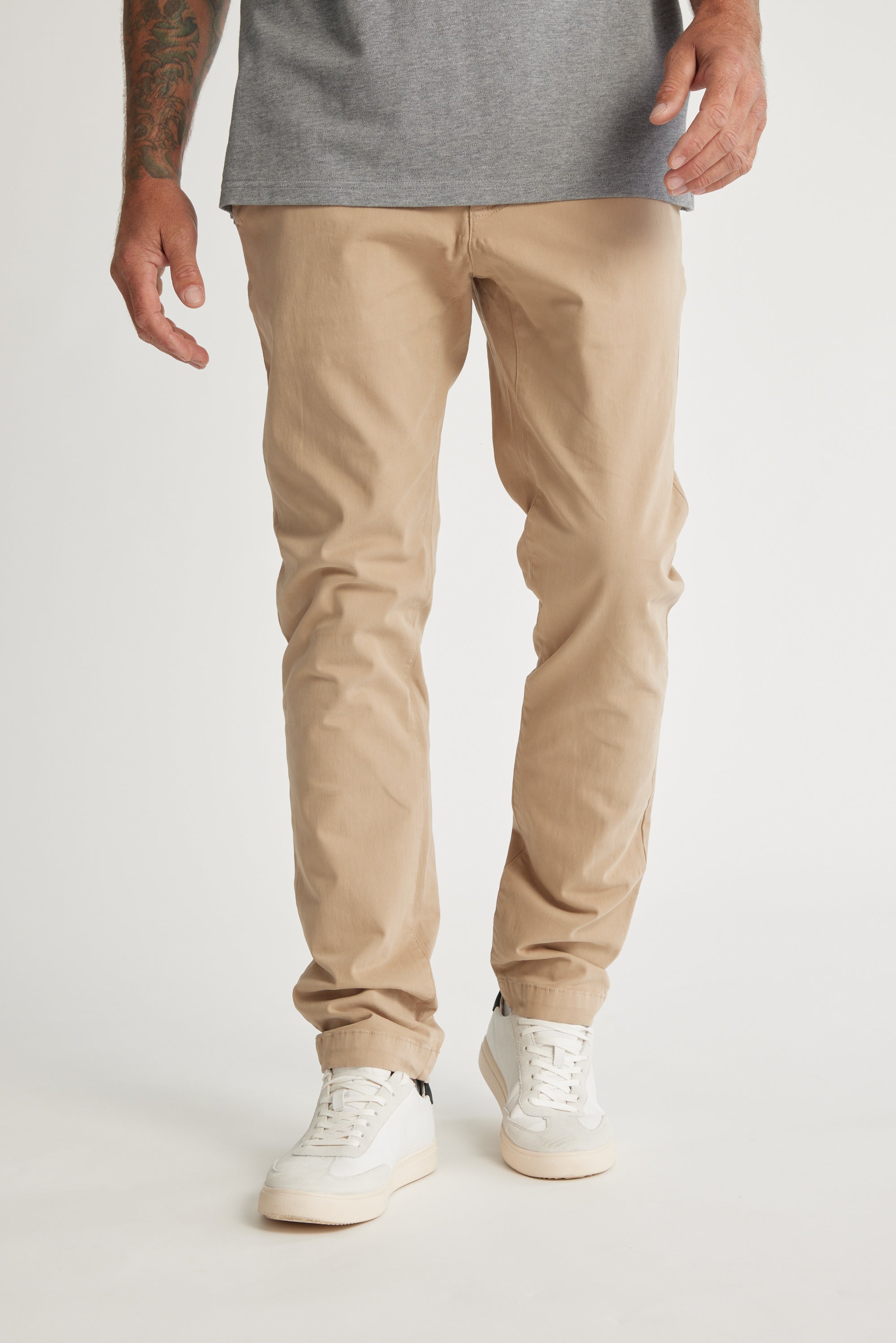 A person, visible only from the waist down, wears Barkers Beauford Chino O men's pants in a comfy cotton-elastane blend with a gray shirt and white sneakers, standing against a plain white background.