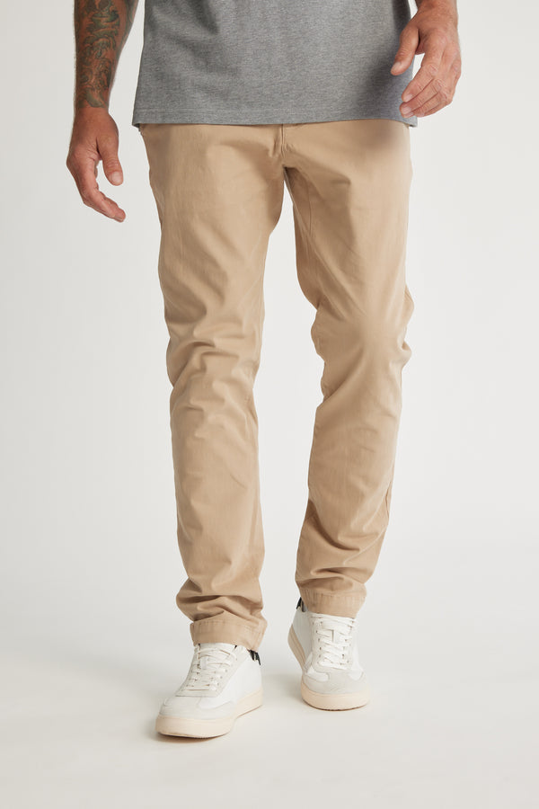 A person, visible only from the waist down, wears Barkers Beauford Chino O men's pants in a comfy cotton-elastane blend with a gray shirt and white sneakers, standing against a plain white background.