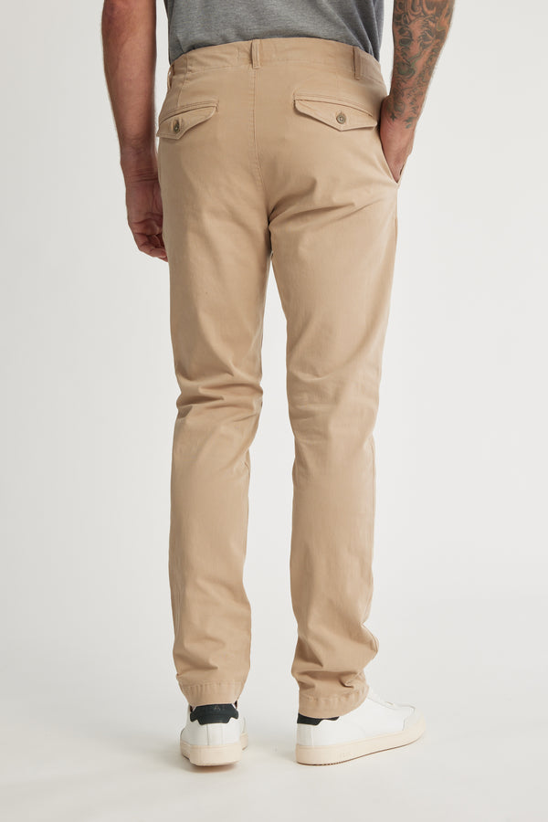 A person stands against a white background, wearing Barkers Beauford Chino O men's pants in beige—a cotton elastane blend—with a gray shirt tucked in, white sneakers, and a tattooed left forearm visible from the back.