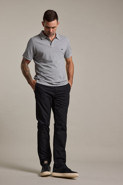 A man stands with his hands in his pockets, looking down. He wears a light gray polo, Barkers Dundas Chino trousers in a relaxed fit, and black shoes against a plain neutral background.