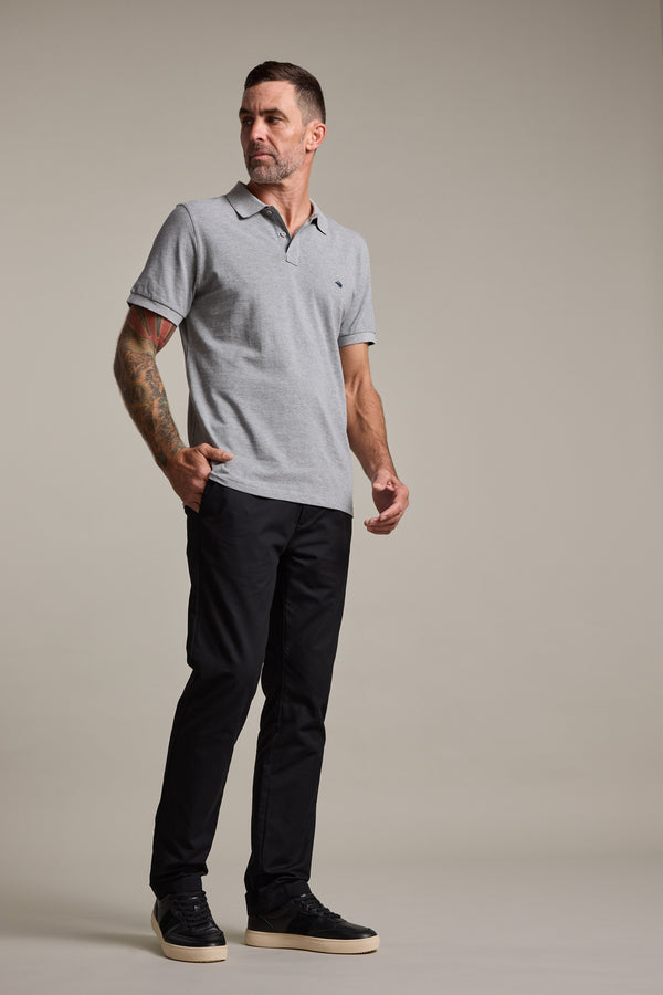 A man with short hair and a tattoo on his right arm stands against a plain background, wearing a light gray polo shirt, Barkers Dundas Chino made from responsibly sourced cotton, and black sneakers with white soles. He looks slightly to the side.