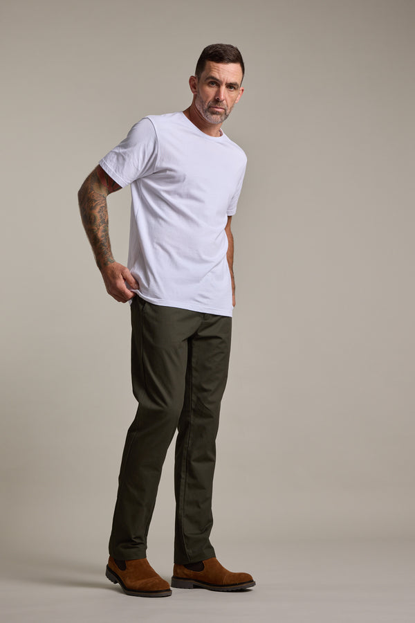 A man with short dark hair and tattoos on his right arm stands against a plain background, wearing a white t-shirt, Barkers Dundas Chino trousers in olive green, and brown boots.