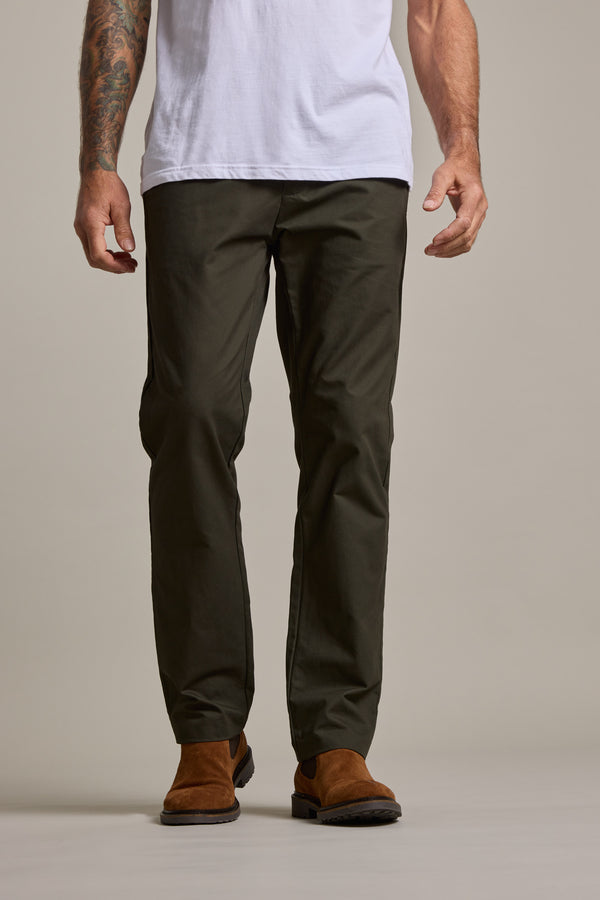 A person wearing Barkers Dundas Chino relaxed fit dark green cotton twill trousers, a white T-shirt, brown suede shoes, and revealing tattooed arms stands on a light-colored floor against a neutral backdrop.