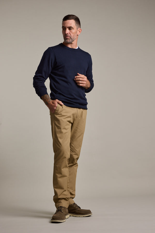 A man in a navy sweater, Barkers Dundas Chino trousers in khaki, and brown shoes stands against a neutral background, looking to the side with one hand near his chest.