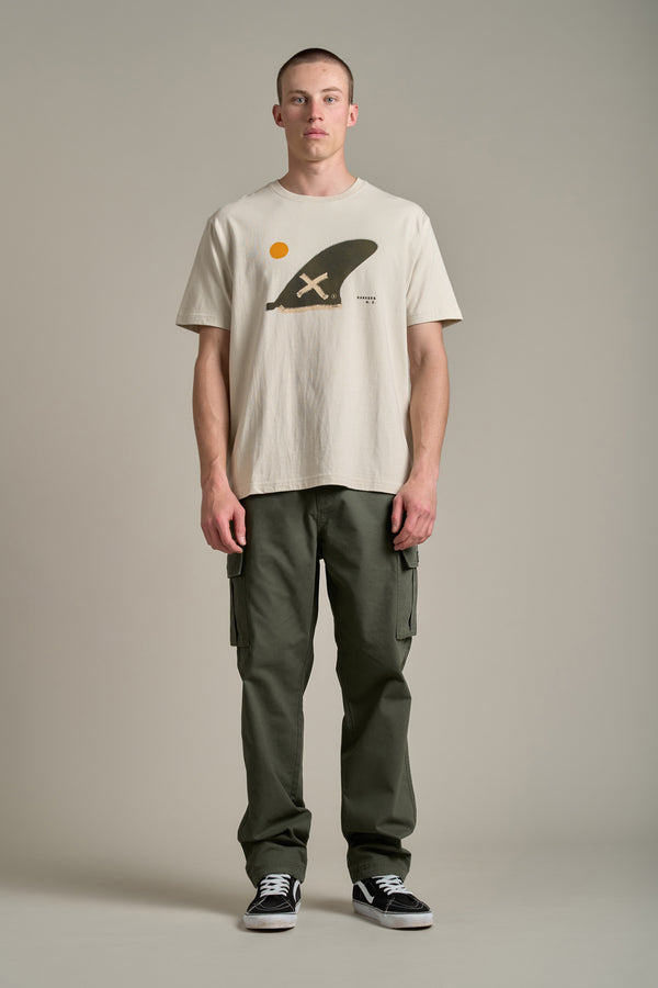 A man stands against a plain background wearing a beige graphic helmet tee, Barkers Randall Cargo Pants in olive green twill cotton for utilitarian style, and black sneakers. He faces forward with a neutral expression and hands at his sides.