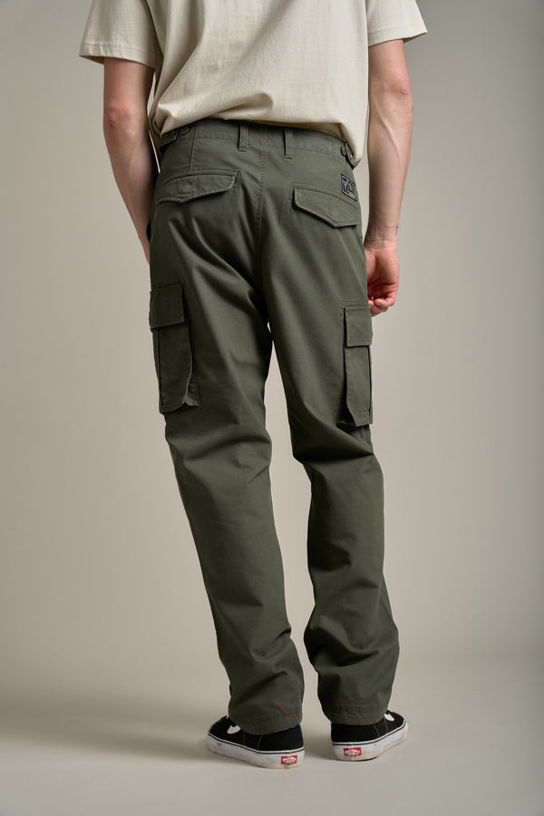 A person stands facing away from the camera, wearing a beige t-shirt, Barkers Randall Cargo Pants in olive green twill cotton with flap pockets, and black-and-white sneakers, set against a neutral background.