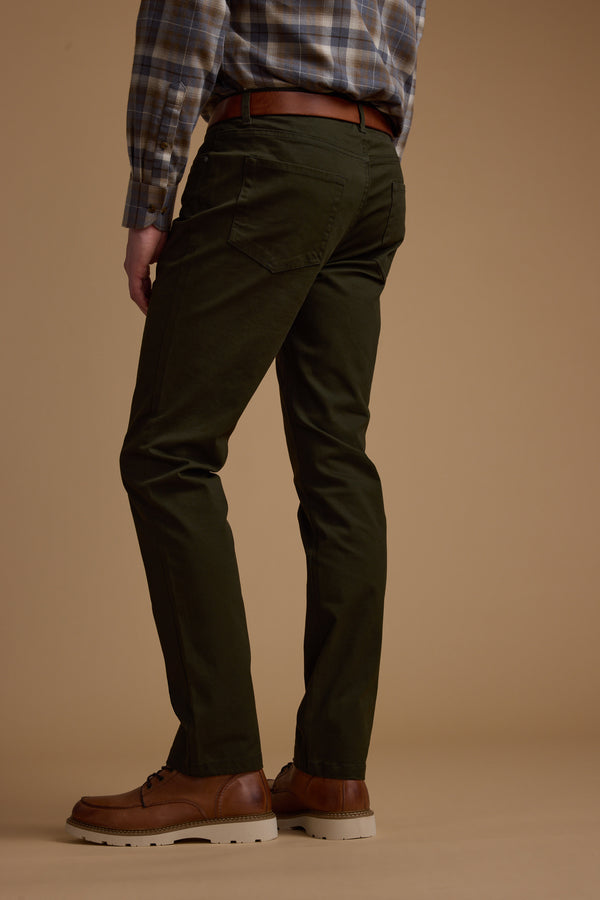 A person stands against a brown background, shown from the back with only the lower body visible, wearing Barkers Beckett 5-Pocket Pant in olive green, paired with a brown belt, plaid shirt, and brown dress shoes.