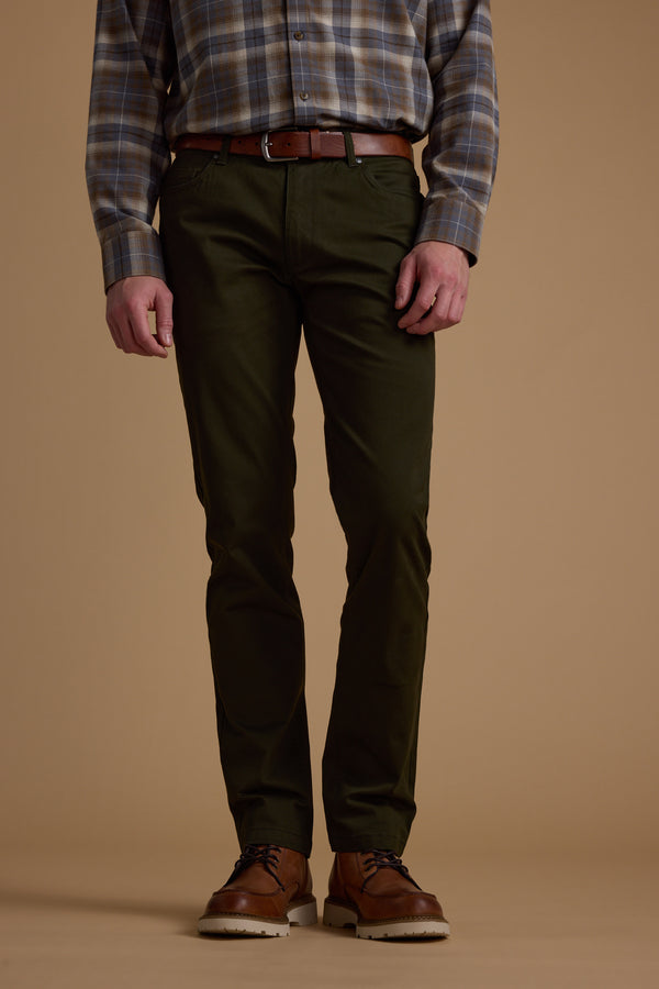 A person stands against a beige background wearing Barkers Beckett 5-Pocket Pant in dark green, paired with a plaid shirt, brown belt, and brown leather shoes. The photo is cropped from the shoulders down.