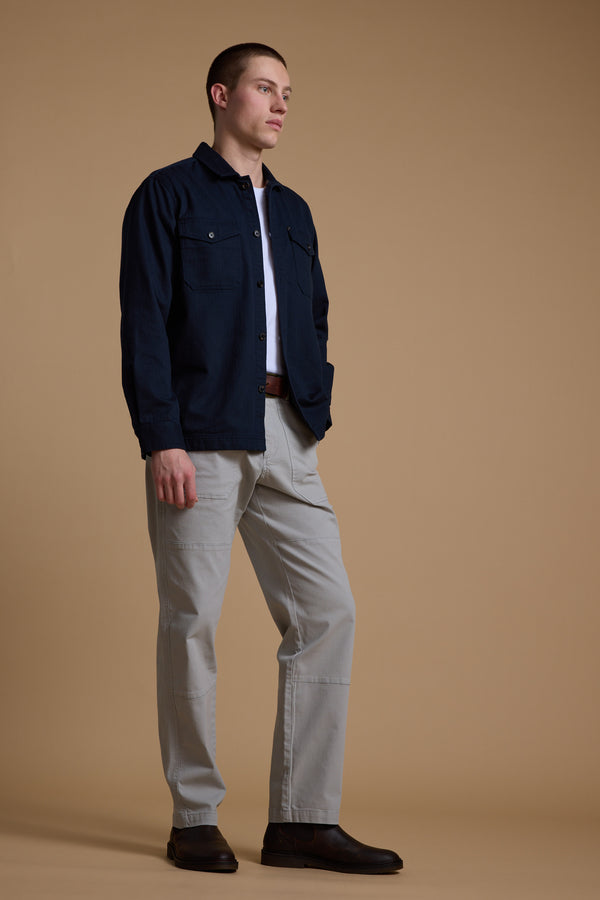 A person stands against a beige background, wearing Barkers' Gifford Herringbone Jacket in dark navy with flannel lining, paired with a white shirt, light gray pants, and dark brown shoes. They look slightly to the side with hands at their sides.