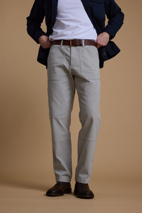 A person, head not visible, stands against a beige background wearing Barkers’ Tidal GD Utility Pant in light grey with a tapered fit, paired with a white shirt, dark jacket, brown belt, and dark brown shoes. Hands are in their pockets.