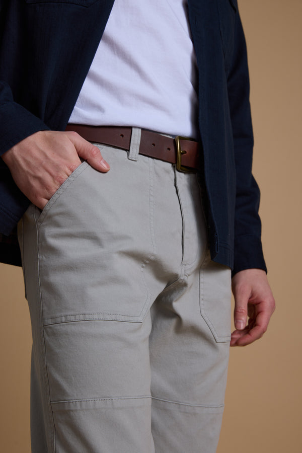 A person wears a white t-shirt, navy jacket, Barkers Tidal GD Utility Pant in light gray, and a brown belt with one hand in their pocket, shown from shoulders to thighs against a plain beige background.