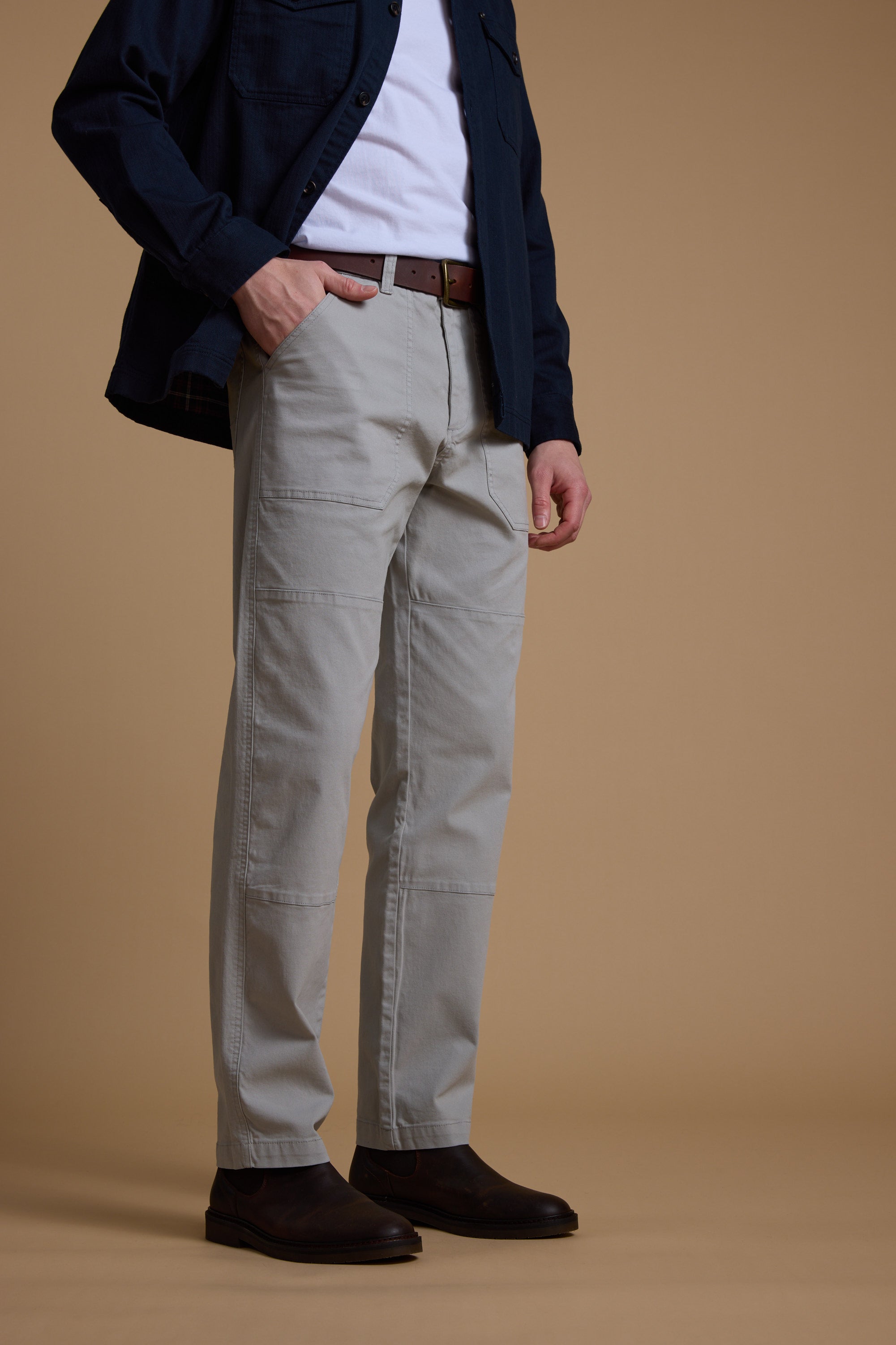 A person stands against a beige background wearing Barkers’ Tidal GD Utility Pant in light gray cotton, paired with a white t-shirt, dark blue button-up shirt, brown belt, and dark brown shoes. The person’s face is not visible.