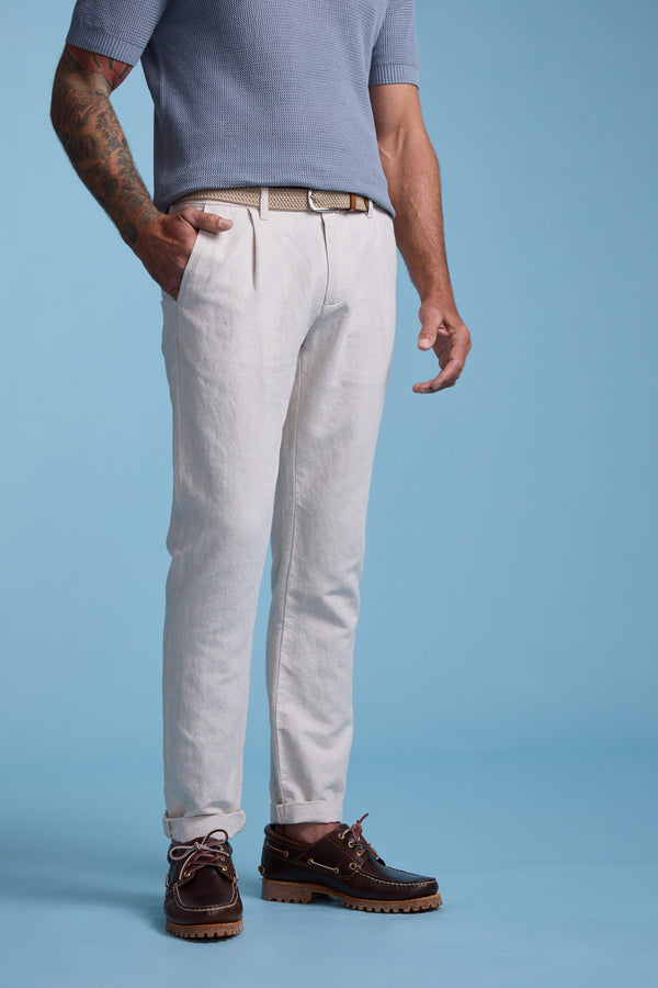 A man stands against a blue background wearing Barkers' Newman Linen Pant in light beige, a short-sleeve blue shirt, tan belt, and dark brown boat shoes. One hand is in his pocket and tattoos decorate his left arm.