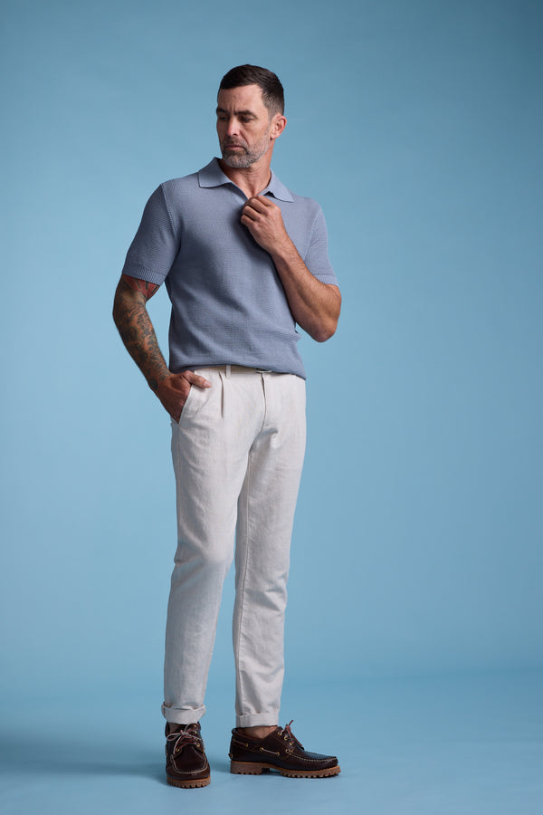 A man with short dark hair and a beard stands against a blue background, wearing a light blue short-sleeve polo, Barkers Newman Linen Pant in light beige, and brown shoes. One hand is in his pocket while the other adjusts his collar.