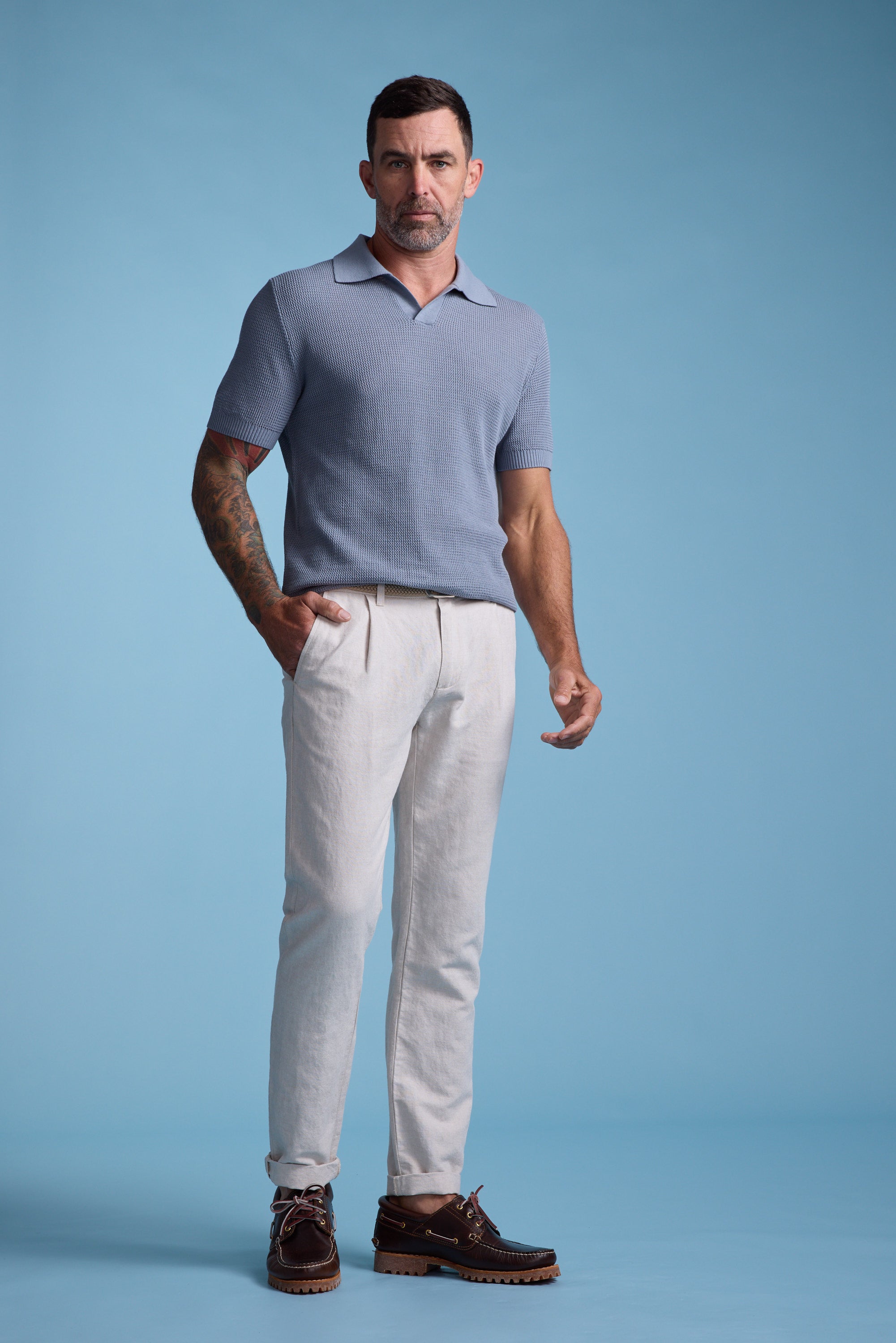 A man with short dark hair and tattoos stands against a blue background, wearing a light blue short-sleeve knit polo, Barkers' Newman Linen Pant in beige, and brown leather shoes.