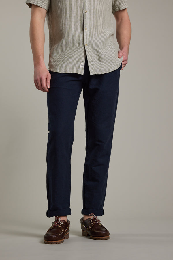 A person stands against a plain background, wearing Barkers' Newman Linen Pant in dark blue with rolled cuffs, a beige short-sleeve button-up shirt, and brown leather shoes. The person's face is not visible.