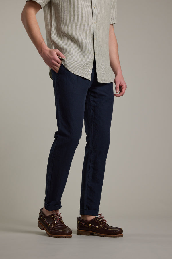 A person stands with one hand in their pocket, wearing the Barkers Newman Linen Pant in dark blue, paired with a short-sleeve, light-colored button-up shirt and brown boat shoes against a plain background.