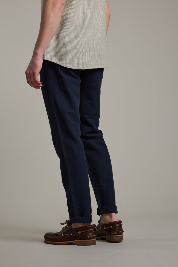 A person stands sideways, wearing a light short-sleeve shirt, Barkers Newman Linen Pant in dark blue, and brown boat shoes against a plain light background.