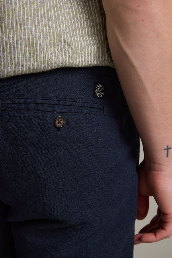 Close-up of a person wearing Barkers' Newman Linen Pant in navy blue with relaxed fit, buttoned back pocket, and black patch. The person also wears a beige striped shirt and has a cross tattoo on their wrist.