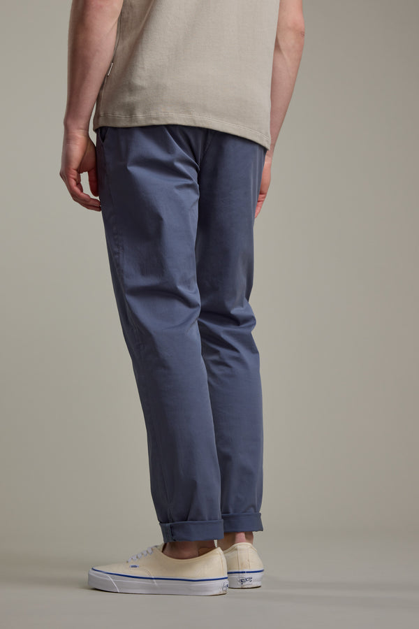 A person stands facing away, wearing a beige shirt, Barkers Kingly Chino Pant in blue, and white sneakers with blue accents. Their lower back, legs, and feet are visible against a plain neutral background.