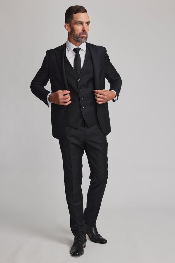 A man with short dark hair and a beard adjusts his Barkers Mayfair ZQrx Waistcoat over a white shirt and black tie, standing against a plain light background while looking off to the side.