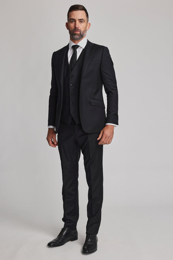 A man with short dark hair and a beard stands facing forward, wearing the Barkers Mayfair ZQrx Waistcoat over a white shirt, black tie, and merino dress shoes against a plain light background.