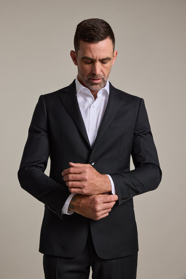 A man wearing the Barkers Mayfair ZQrx Suit Jacket and a white dress shirt stands against a plain background, looking down as he adjusts his left cuff with his right hand.