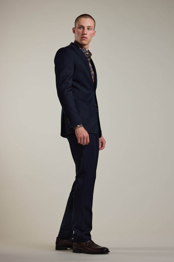 A man in a dark suit with a patterned shirt stands sideways, looking over his shoulder against a plain light background. His Barkers Croydon Twill Suit Trouser adds refinement and comfort to his modern look.