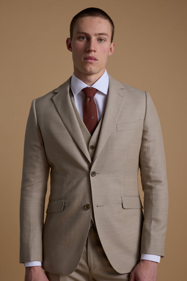 A man in the Barkers Lister Texture Suit Jacket, featuring a beige wool-linen blend, paired with a white shirt and brown tie, stands against a tan backdrop—an ideal summer wedding suit.
