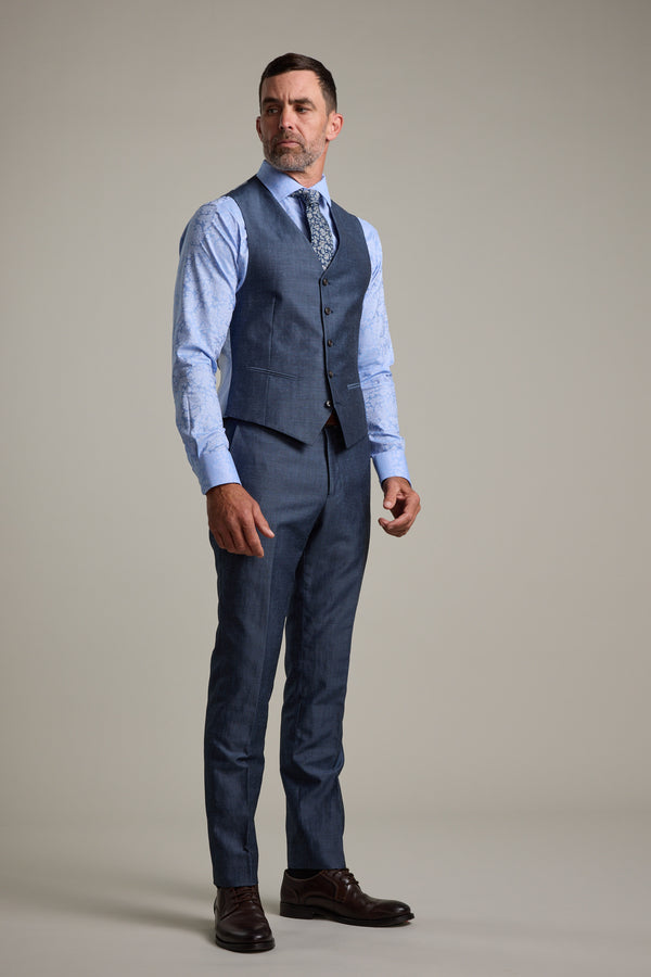 A man stands against a plain backdrop, wearing the Barkers Charles Texture Waistcoat with an adjustable back buckle, part of a blue three-piece suit. He pairs it with a patterned tie and dark brown dress shoes, looking slightly to the side with a serious expression.