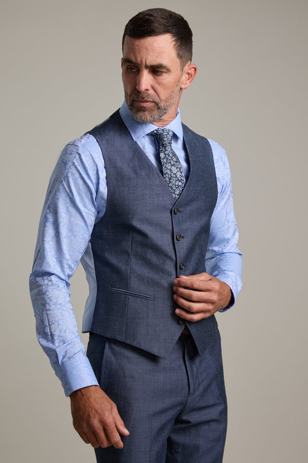 A man with short dark hair and a beard wears a blue patterned shirt and the Barkers Charles Texture Waistcoat with matching suit pants, adjusting his vest while looking left. The background is plain and light-colored.