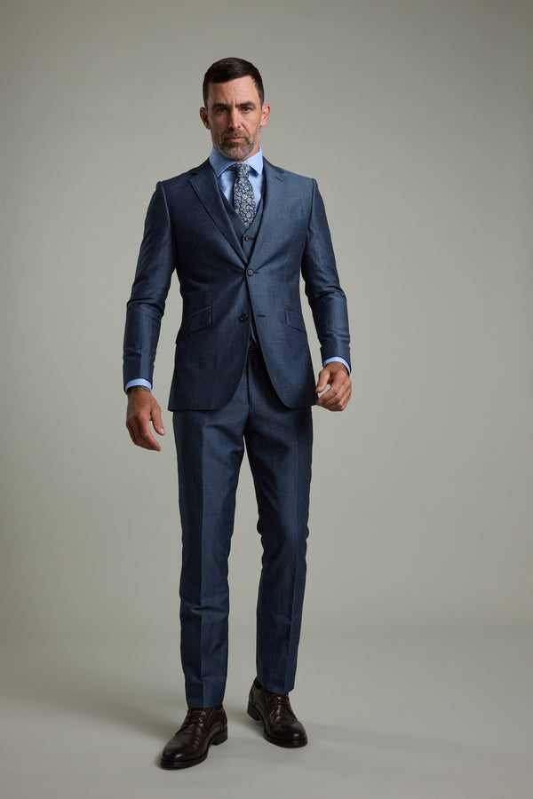 A man faces forward wearing Barkers’ Charles Texture Suit Trouser in steel grey, paired with a light blue shirt, patterned tie, dark brown dress shoes, and posed against a plain light grey background.