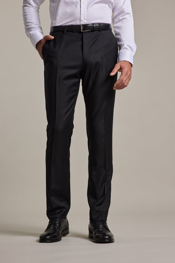 A man stands against a plain background with one hand in his pocket, dressed in a white shirt, black belt, Mayfair ZQrx Suit Trouser by Barkers, and black dress shoes.