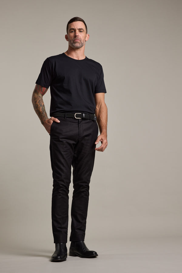 A man with short dark hair, a beard, and a tattoo on his left forearm stands against a plain background in a black t-shirt and Barkers Lyndon Cotton Pants, styled smart-casual with a black belt and boots and wearing a neutral expression.