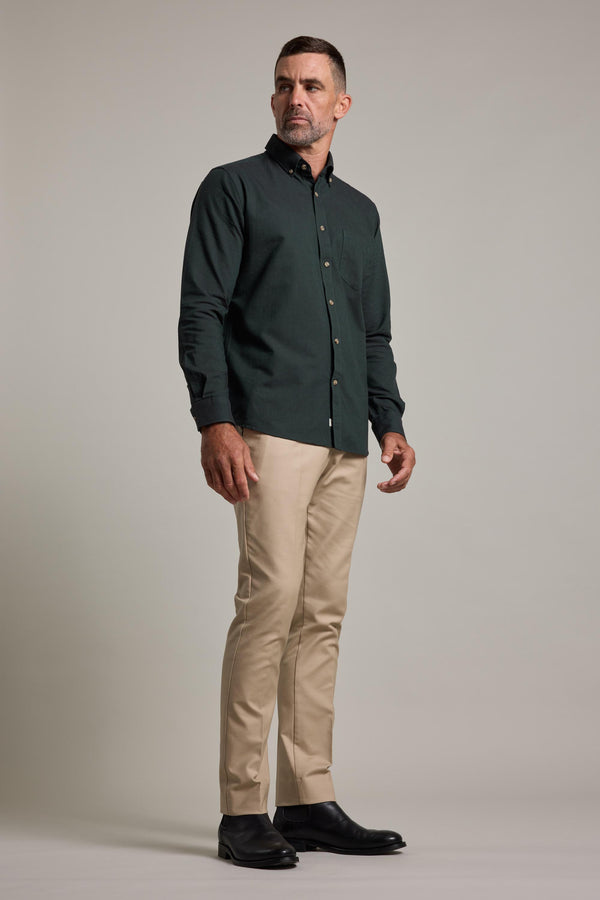 A man with short dark hair and a trimmed beard stands against a plain background wearing the Barkers Thatcher Oxford Shirt in dark green, tailored fit, paired with beige pants and black shoes, looking slightly to his left.