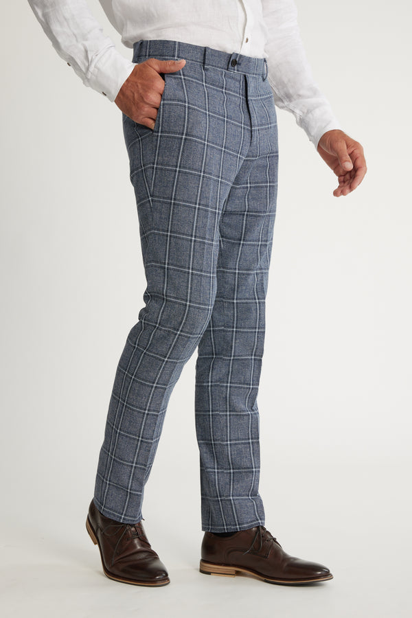 A man wearing Barkers Highland Check Suit Pant, a white long-sleeve shirt, and brown leather dress shoes stands with one hand in his pocket against a plain white background.